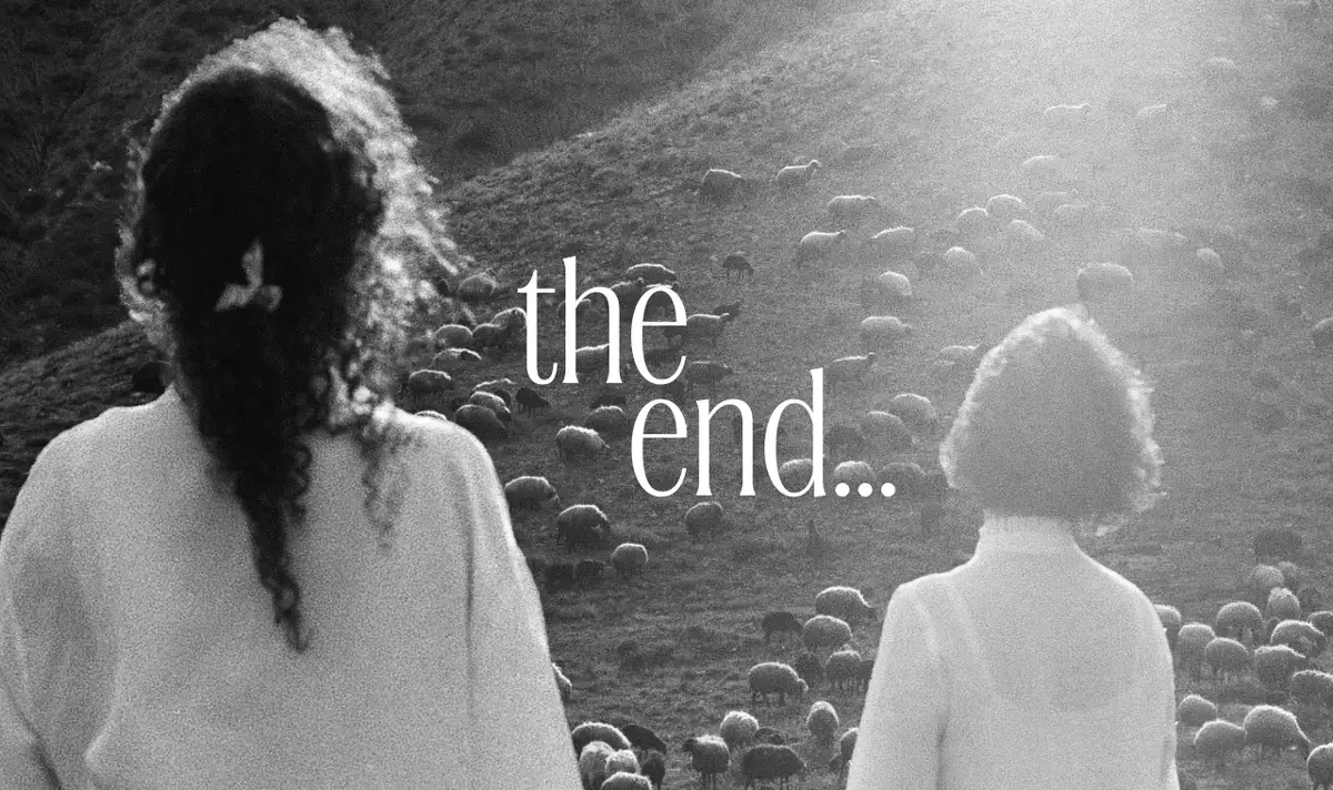Two figures viewed from behind facing a hillside covered in sheep, with "the end..." text overlaid in white, in black and white cinematographic style
