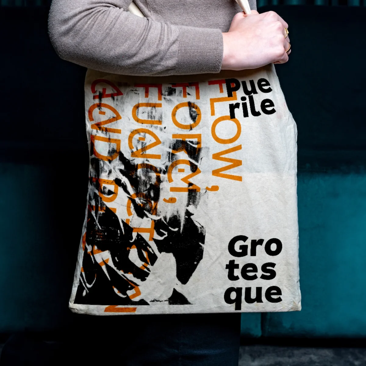 White canvas tote bag with overlapping orange and black typography reading "Puerile Grotesque" held by a person wearing a grey jumper