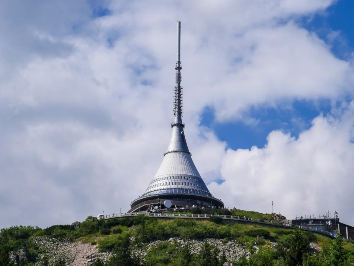 Hotel Ještěd perches atop a forested hill, its distinctive white conical tower and antenna rising against a blue sky with white clouds