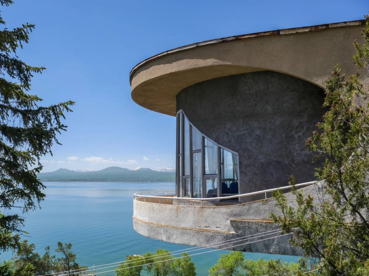Circular concrete structure with curved overhanging roof and floor-to-ceiling windows perched on a hillside above a turquoise lake, surrounded by evergreen trees