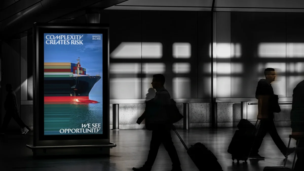 Commuters walk past a backlit advertising display showing a cargo ship with the text "Complexity creates risk" and "We see opportunity" for White & Case's new brand identity