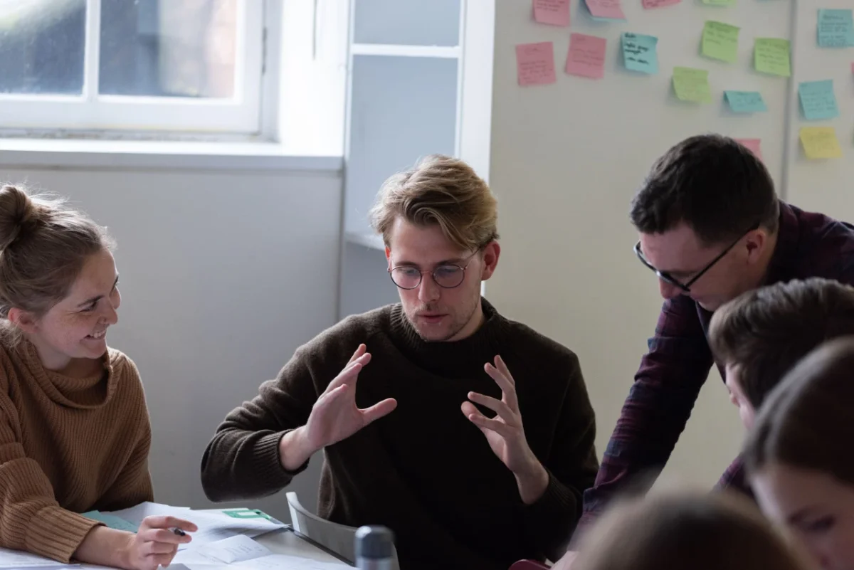 Five people gathered around a table in a bright workshop space with coloured sticky notes on the wall behind them, discussing and collaborating on a project