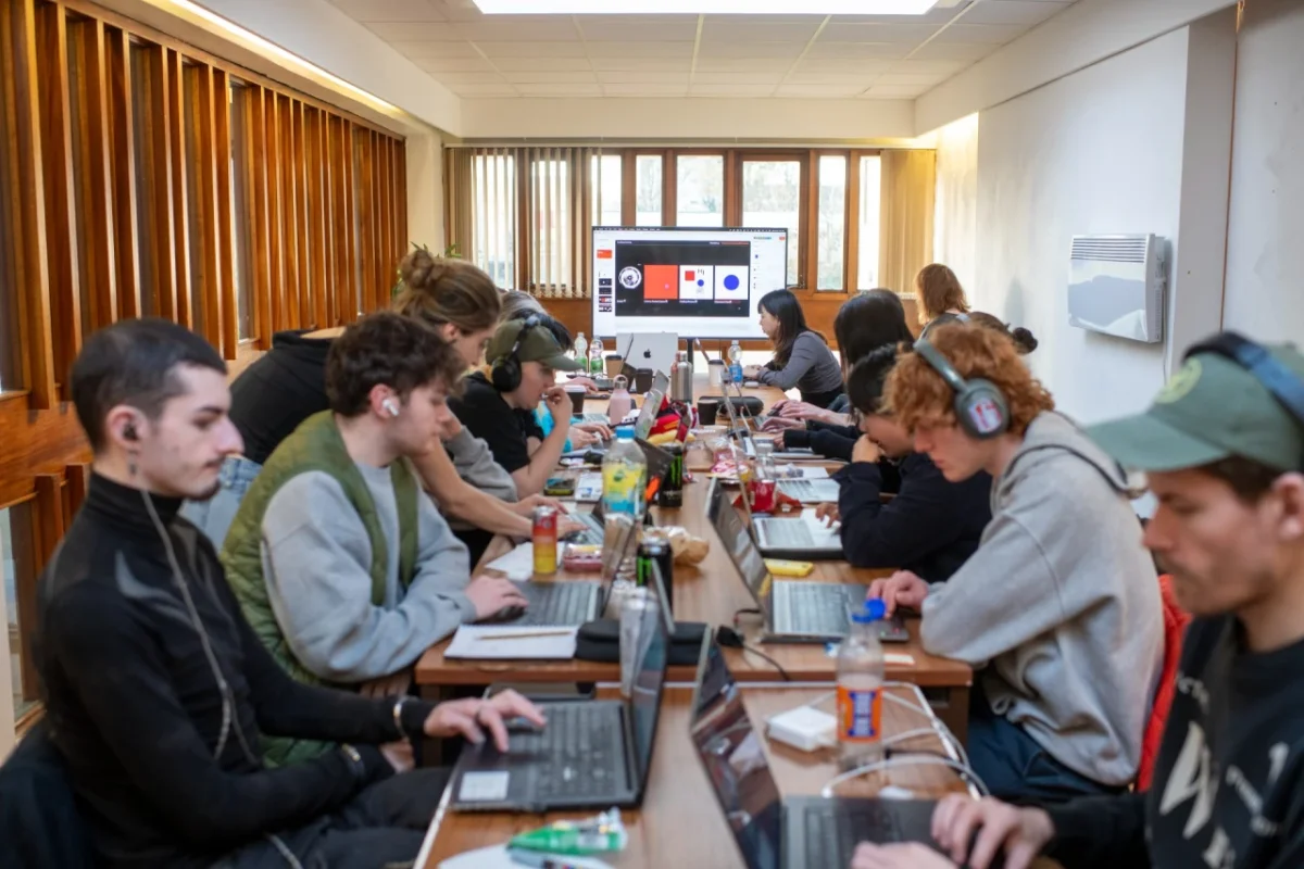 Participants sit at a long table during an International Assembly workshop, working on laptops with refreshments, whilst a screen displays design work in the background