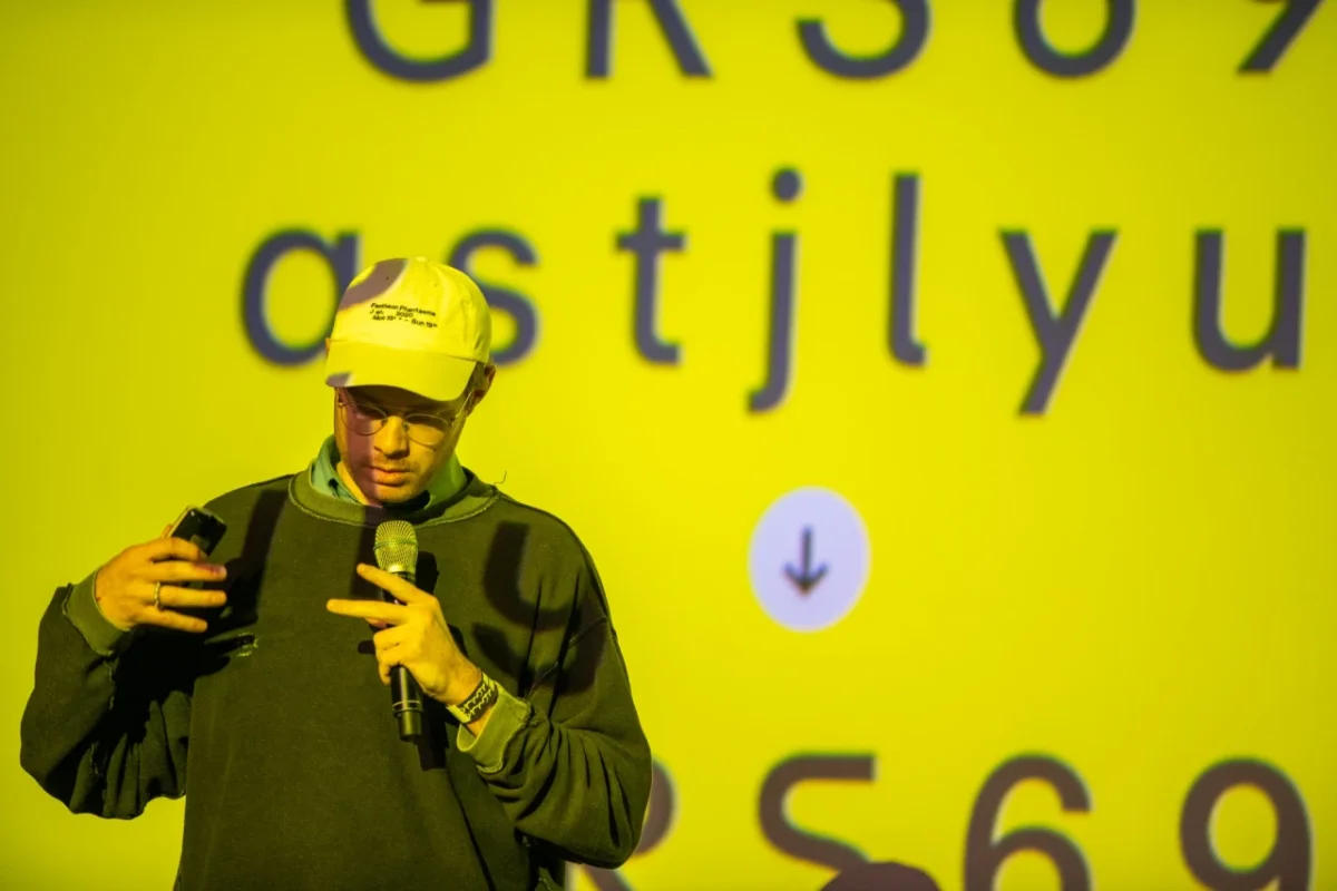 Speaker wearing a yellow cap and olive jacket gestures while addressing an audience in front of a large yellow projection screen at International Assembly