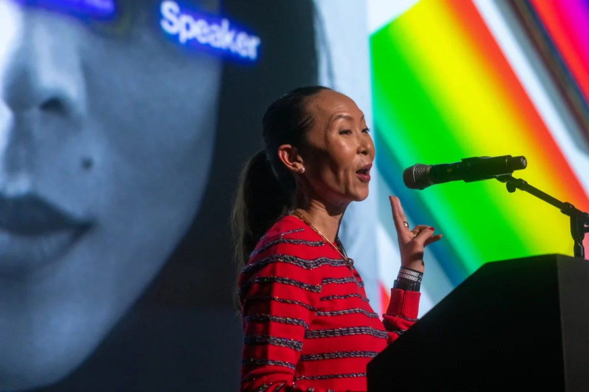 Speaker at a microphone during International Assembly, wearing a red and black striped top in front of a screen displaying a portrait and colourful gradient graphics