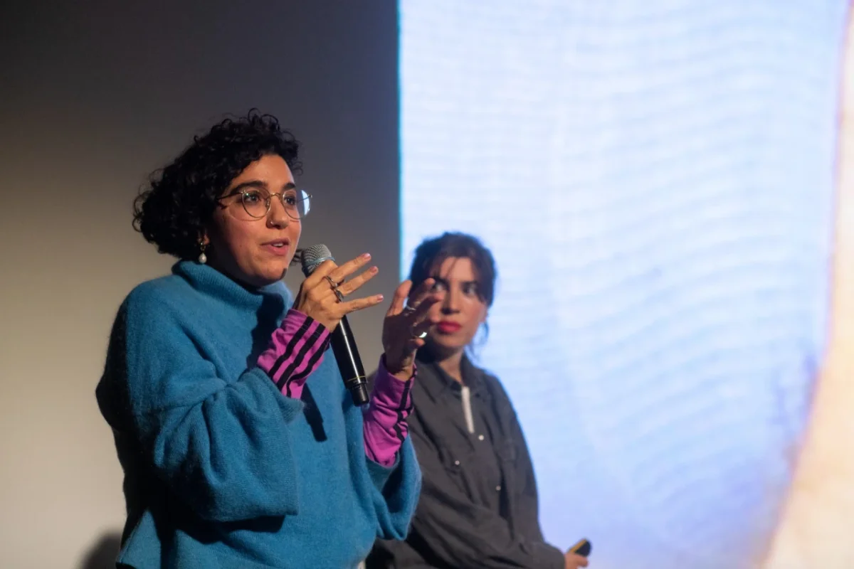 Two speakers gesture whilst presenting on stage at an International Assembly event, with a projection screen visible behind them