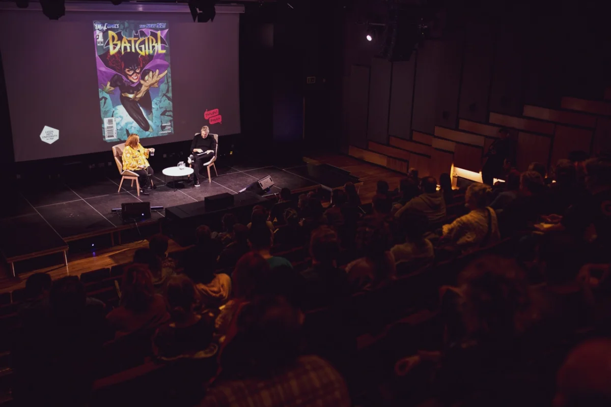 Two people sit on stage during a panel discussion in front of an audience, with a Batgirl comic book cover projected on the screen behind them