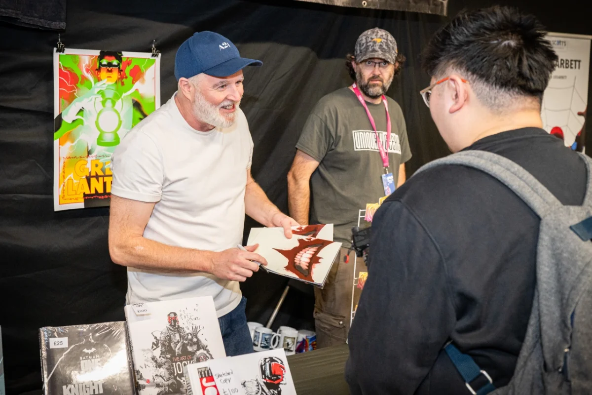 Man in white t-shirt and blue cap signs autographs at a comic convention booth for visitors including a young person in black jacket
