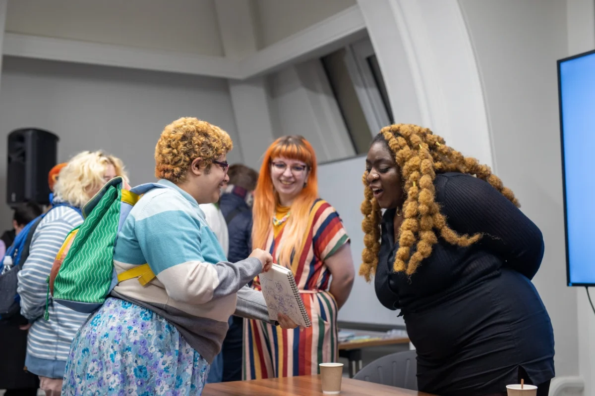 Three people smiling and chatting at an indoor event, including someone with golden curled braids wearing a black top and glasses, and another person with orange fringe and a striped top