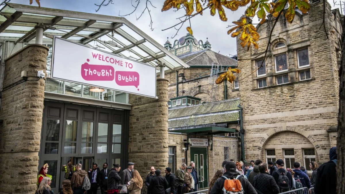 Visitors gather outside Thought Bubble's entrance beneath a pink neon welcome sign, with stone buildings and autumn foliage visible in the background