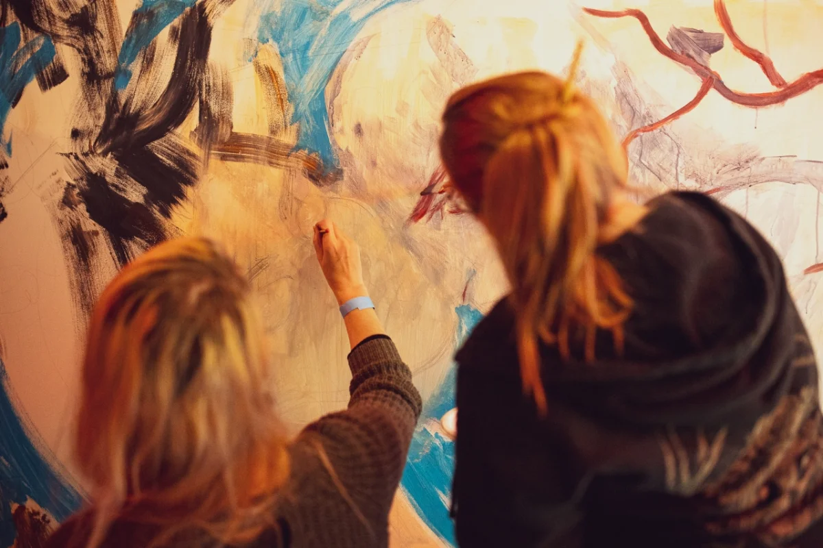 Two people with red hair painting colourful abstract designs on a large canvas wall together
