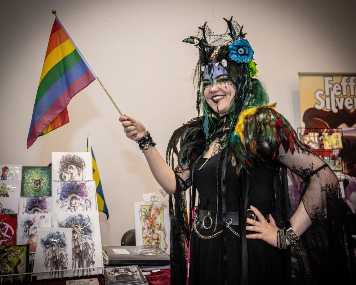 Person in black outfit with feathered wings and blue-tinted hair holding a rainbow pride flag