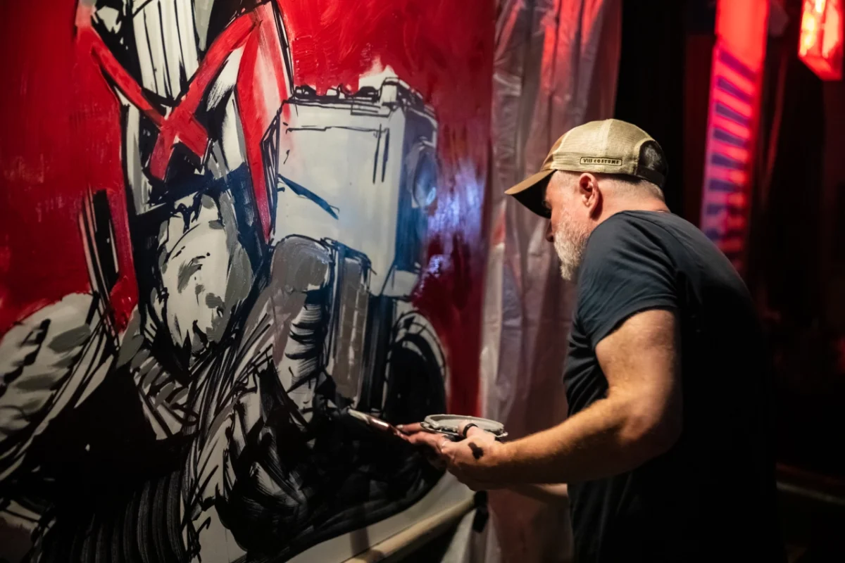 Artist in black cap and t-shirt working on a large-scale black and red mural featuring abstract figures against a vivid red background
