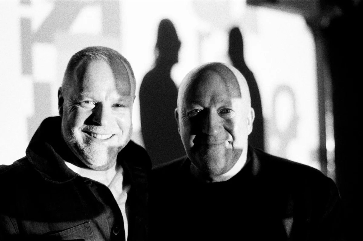 Two men smiling at the camera in black and white photograph with shadows cast on wall behind them