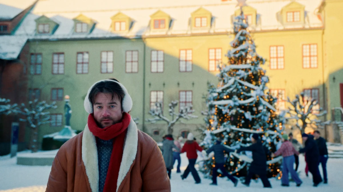 Halfdan stands in a snowy Oslo city square wearing a brown coat and red scarf, with a decorated Christmas tree and colourful historic buildings behind him