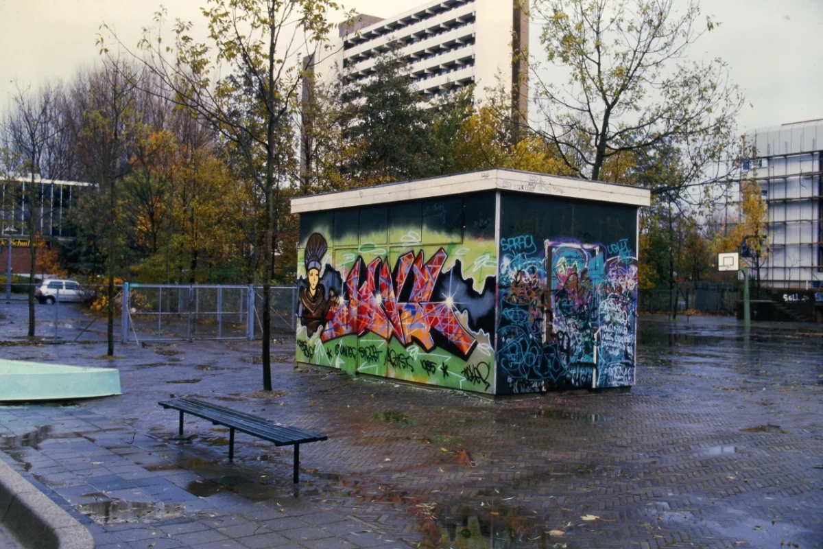 Wet urban playground with a small kiosk covered in vibrant red and blue graffiti art, surrounded by autumn trees and apartment blocks