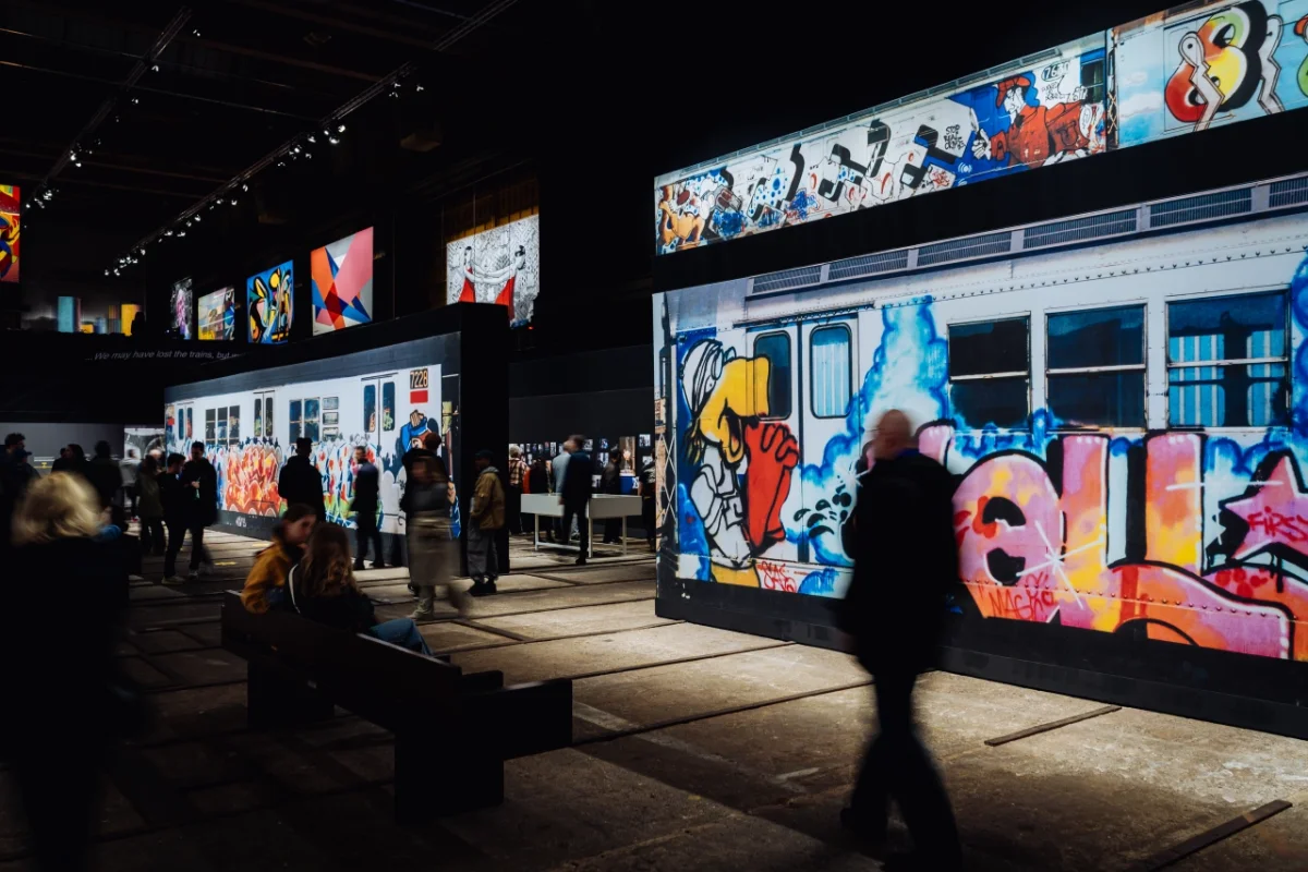 Visitors walk through a darkened gallery space at Straat museum, viewing large illuminated panels featuring vibrant street art and graffiti documentation displayed on walls and suspended screens