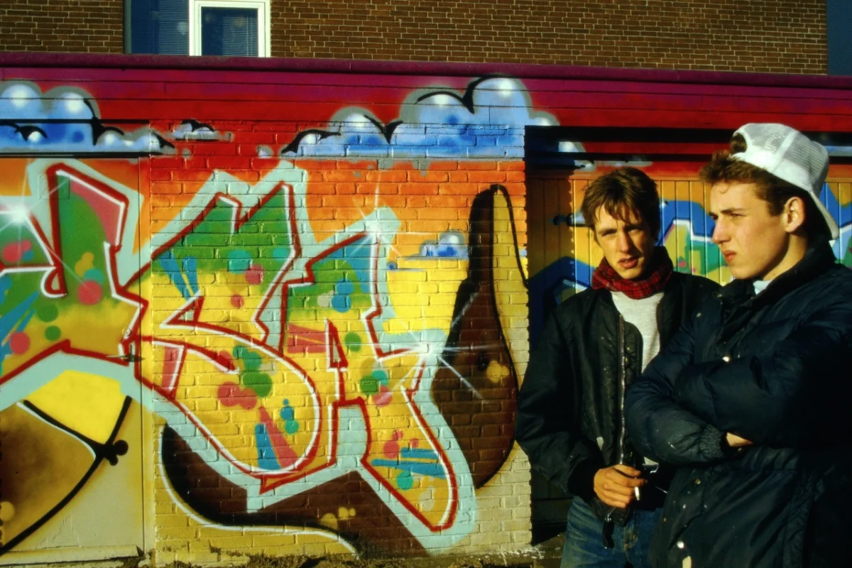 Two graffiti artists stand in front of a vibrant, colourful USA piece with red, yellow, green and blue lettering on a brick wall, Amsterdam 1985