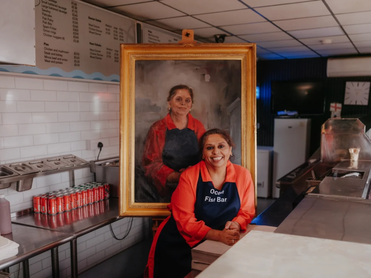 A woman wearing a red jacket and black apron stands in a fish and chip shop kitchen next to her oil portrait in a gilt frame, painted by Royal portrait artist Nicky Philipps
