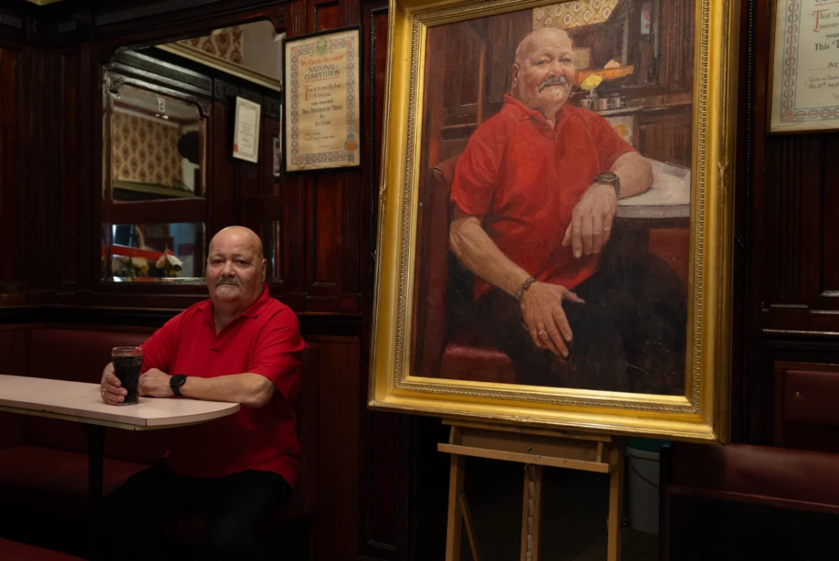 Man in red shirt seated at a table next to his formal oil portrait in a gilt frame, hung on dark wood panelling.