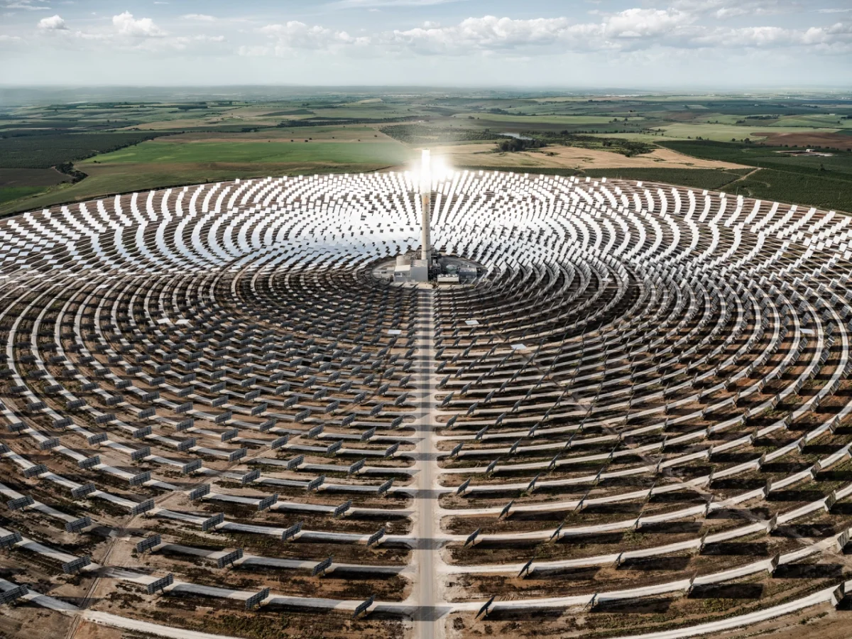 Aerial view of a vast solar thermal power plant with concentric circular rows of mirrors radiating from a central tower beneath cloudy skies, surrounded by agricultural fields