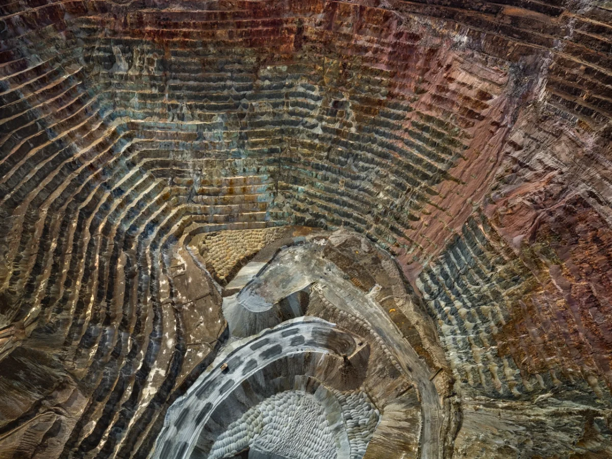 Aerial view of a vast open-pit mine with concentric terraced layers in shades of green, brown and rust, revealing the scale of mineral extraction