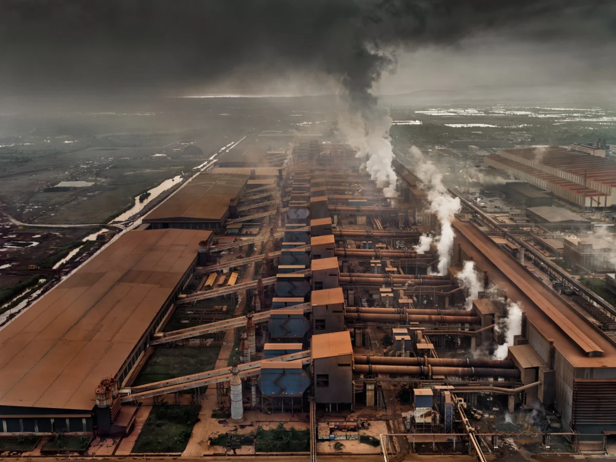 Aerial view of an industrial mining complex with multiple rectangular extraction ponds in orange and blue, surrounded by farmland, with white smoke rising from processing facilities