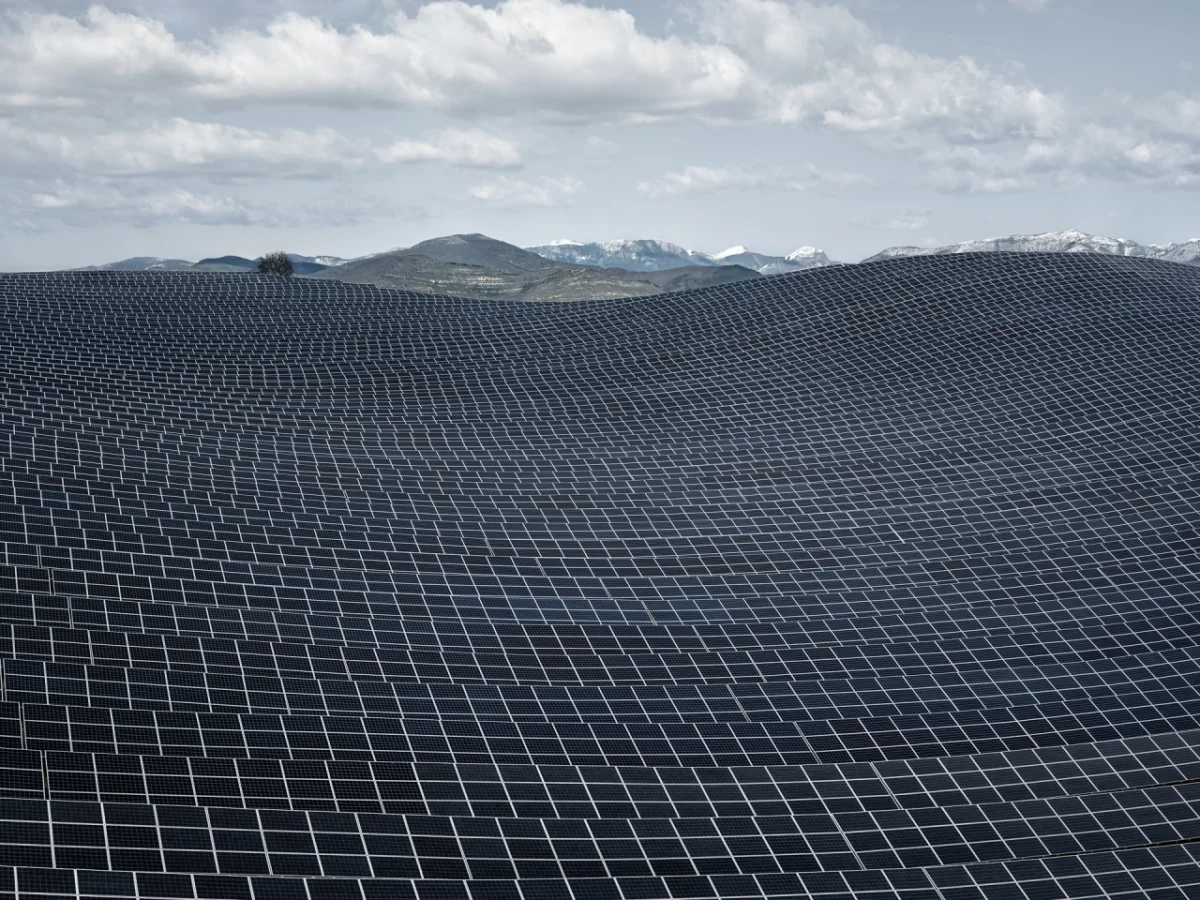 Vast expanse of solar panels stretching across rolling terrain towards snow-capped mountains under a cloudy sky