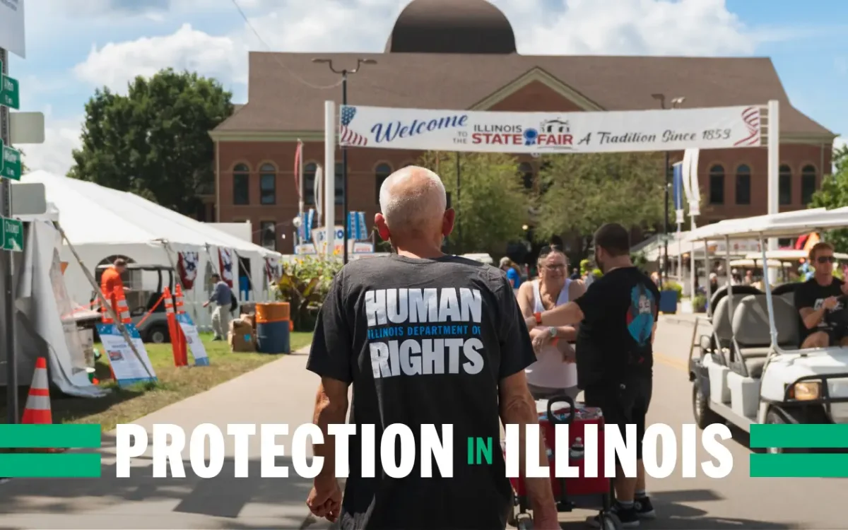 Person wearing a black Human Rights Protection in Illinois t-shirt stands in front of a brick building at an outdoor event with market stalls and visitors
